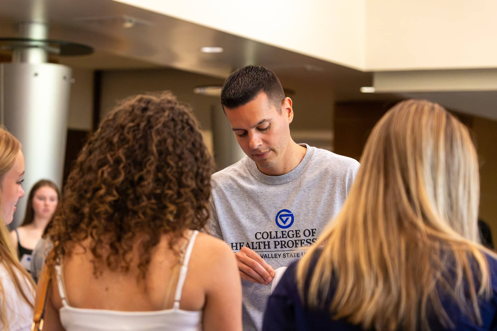 Man in College of Health Professions gray t-shirt hands papers to students in front of him